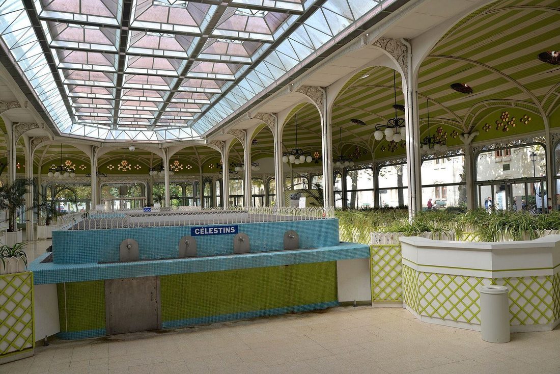 Inside the Hall des Sources in Vichy: a glazed roof on slender white iron columns, green vaulted ceiling panels and the tiled Celestins drinking fountain below.