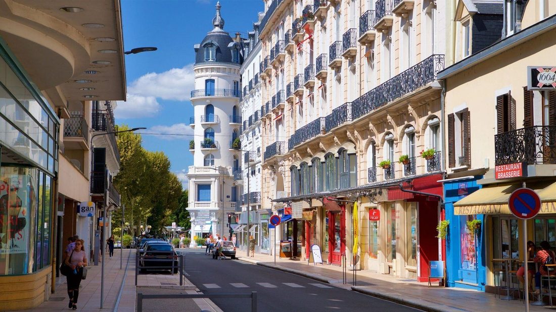 A sunlit street in Vichy: belle-&eacute;poque facades with iron balconies, awnings over the shopfronts, a corner turret and people walking in the shade of the plane trees.
